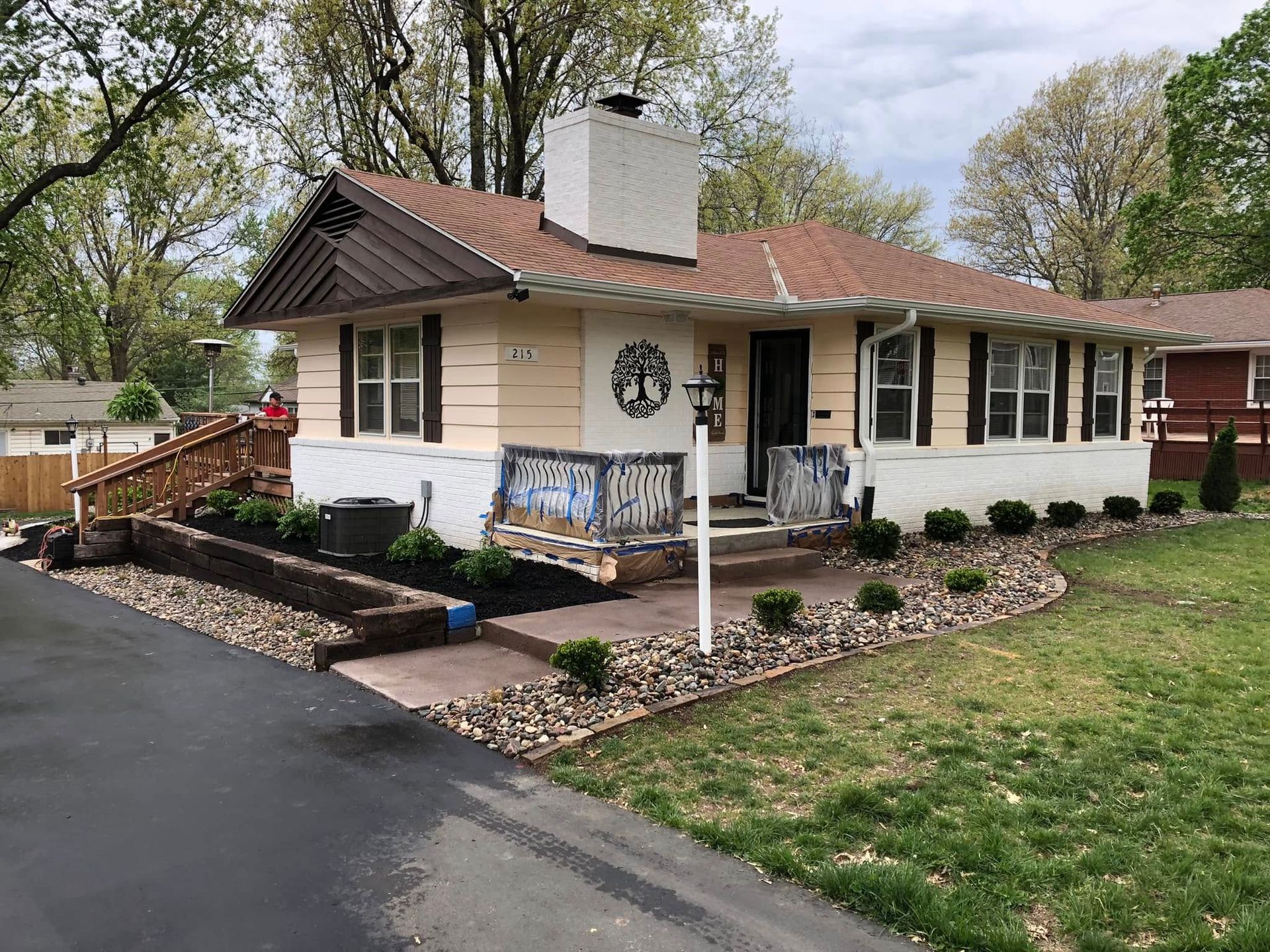 A cream-colored ranch house with a brick chimney, wooden wheelchair ramp, and landscaped rock beds under a cloudy sky.