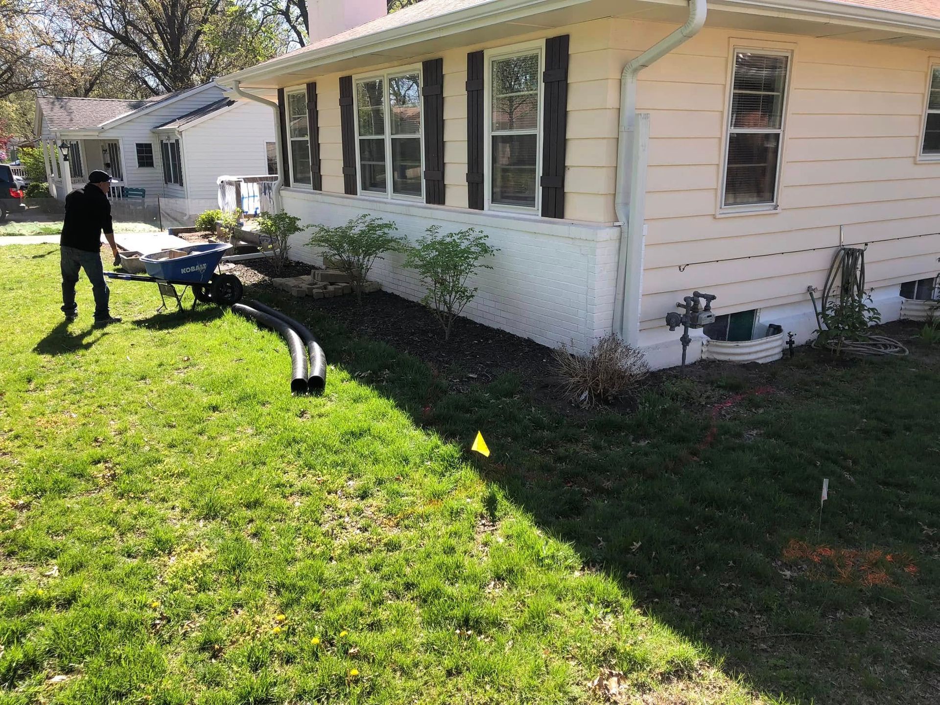 A person stands next to a wheelbarrow and drainage pipes in the yard of a light yellow house with dark window shutters.