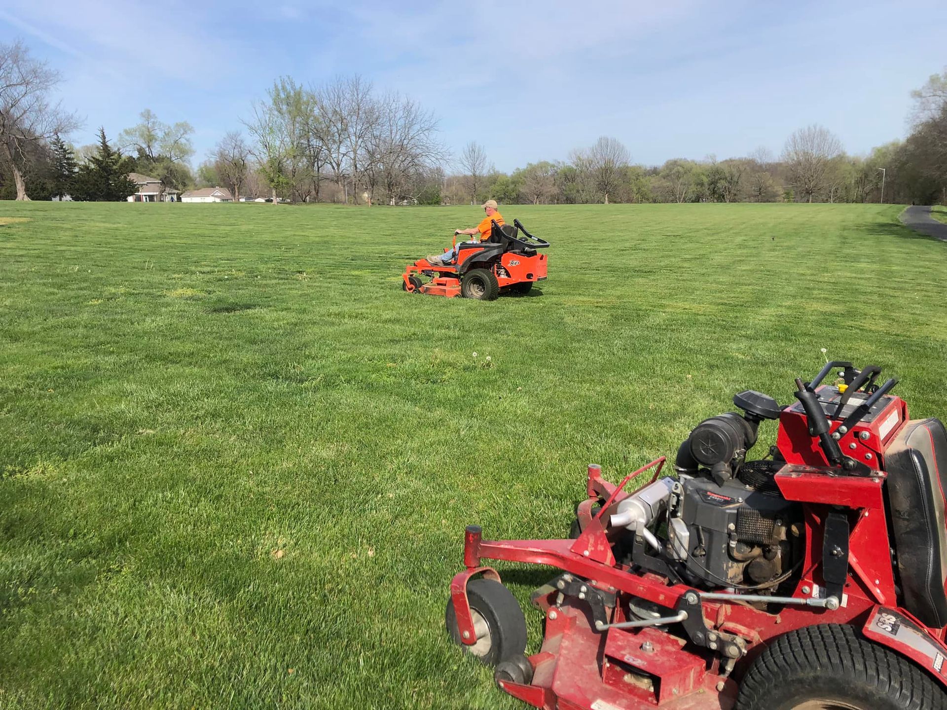 A red zero-turn mower sits in the foreground as an orange mower operates in a large, grassy field under a sunny sky.