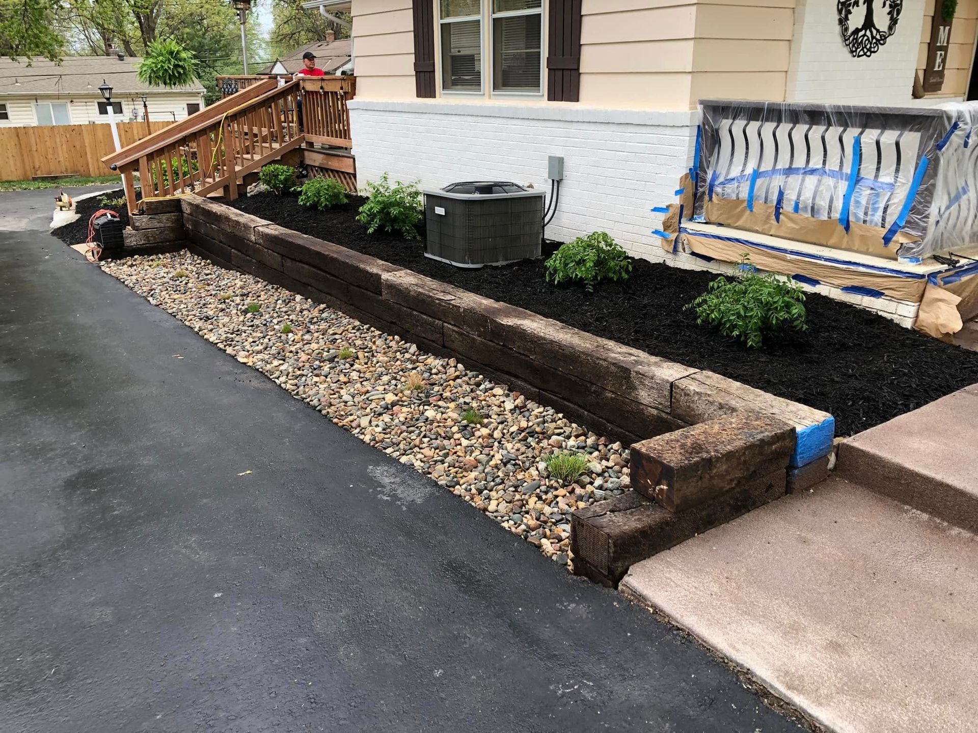 A raised landscape bed with railroad tie walls, black mulch, small green shrubs, and river rock bordering an asphalt driveway