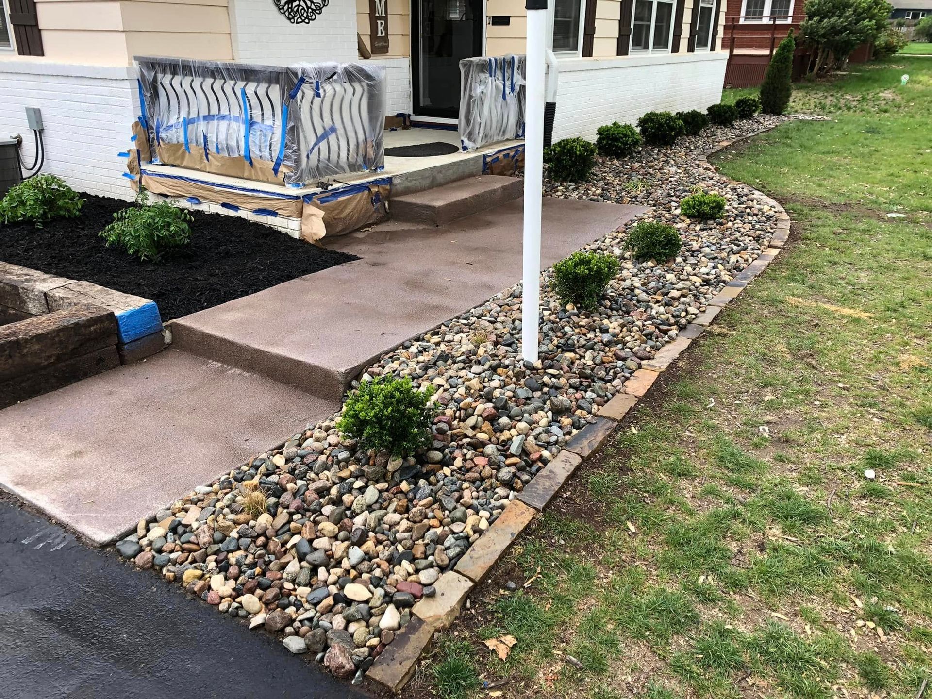 A paved walkway leading to a house entrance, bordered by a rock garden with small shrubs and a dark mulch bed.