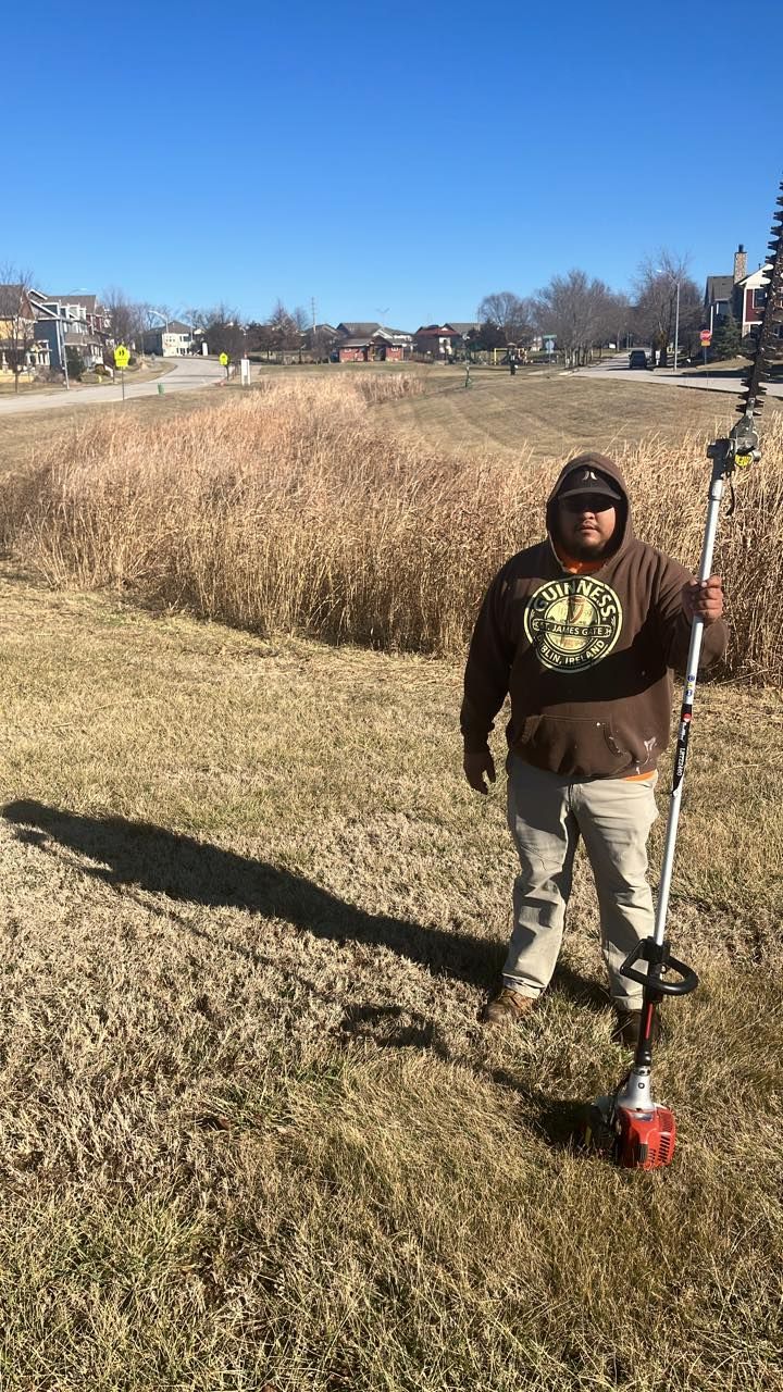 A person in a brown hoodie standing in a field holding a long-handled power hedge trimmer on a sunny day.