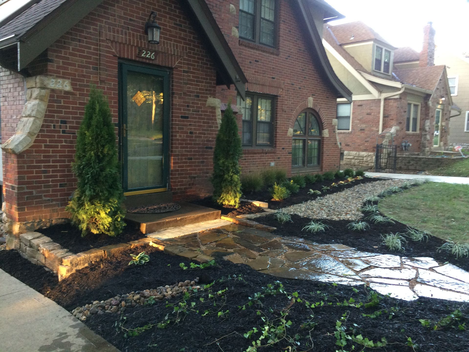 A red brick house with a stone walkway, landscaped garden beds with dark mulch, and two evergreen shrubs by the entrance.