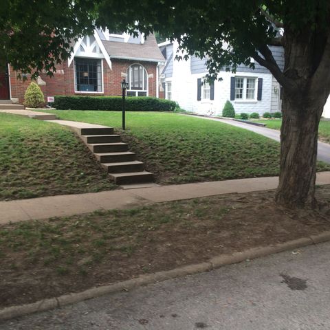 A red brick house with a front staircase leading to the sidewalk, next to a white house with a tree in the foreground.