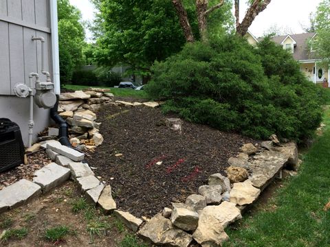 A mulched garden bed next to a house wall, bordered by light-colored stacked stones and featuring a large green shrub.