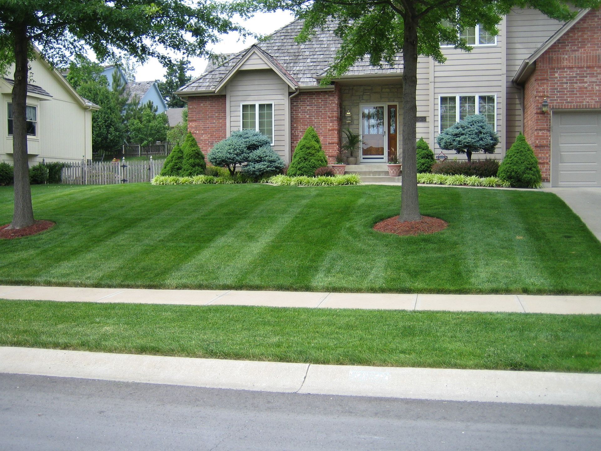 A well-manicured residential lawn with neat mowing stripes, framed by two large trees in front of a brick and siding house.