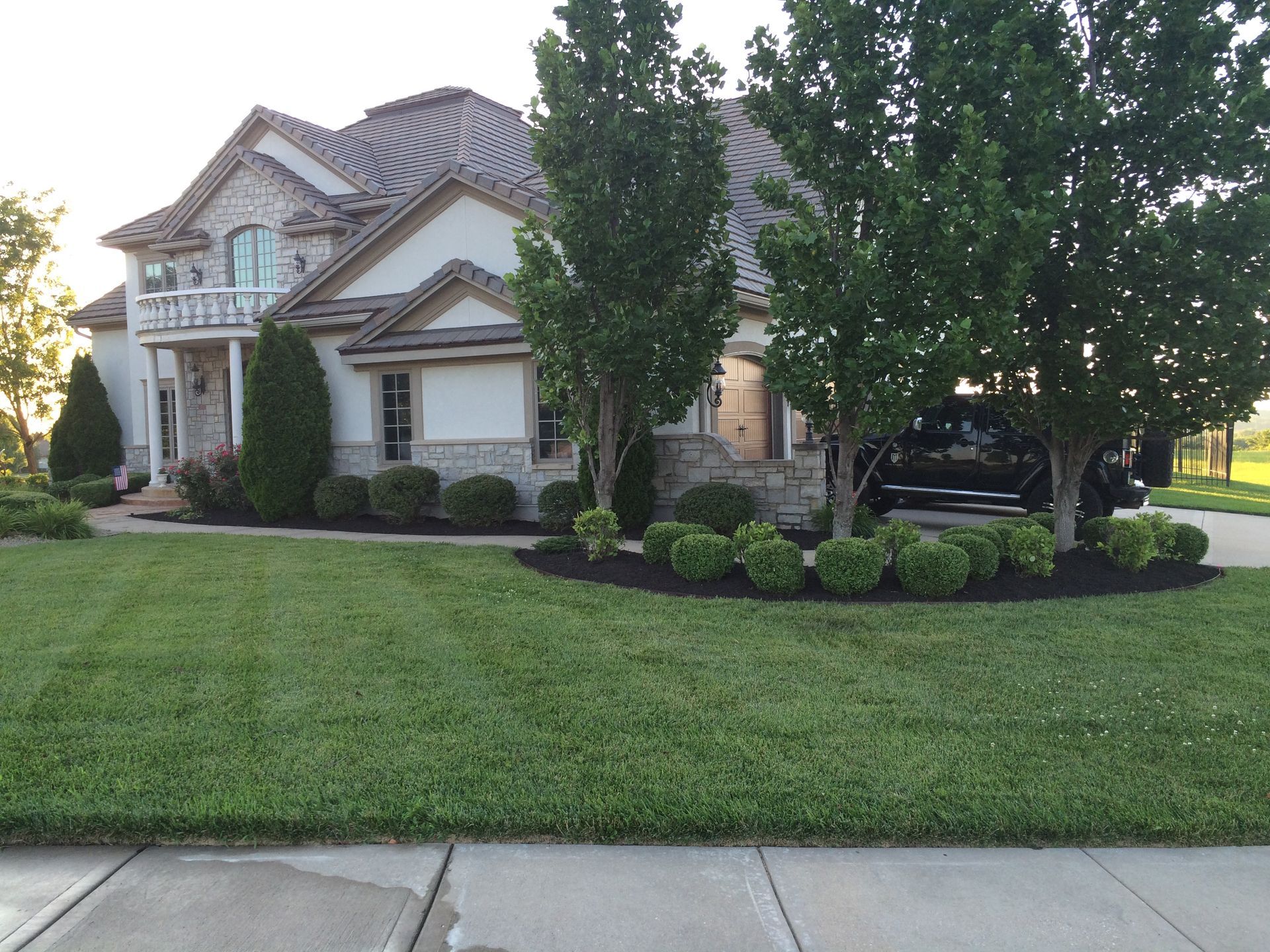 A two-story suburban house with stone and cream siding, a manicured lawn, and landscaping in front of a driveway.
