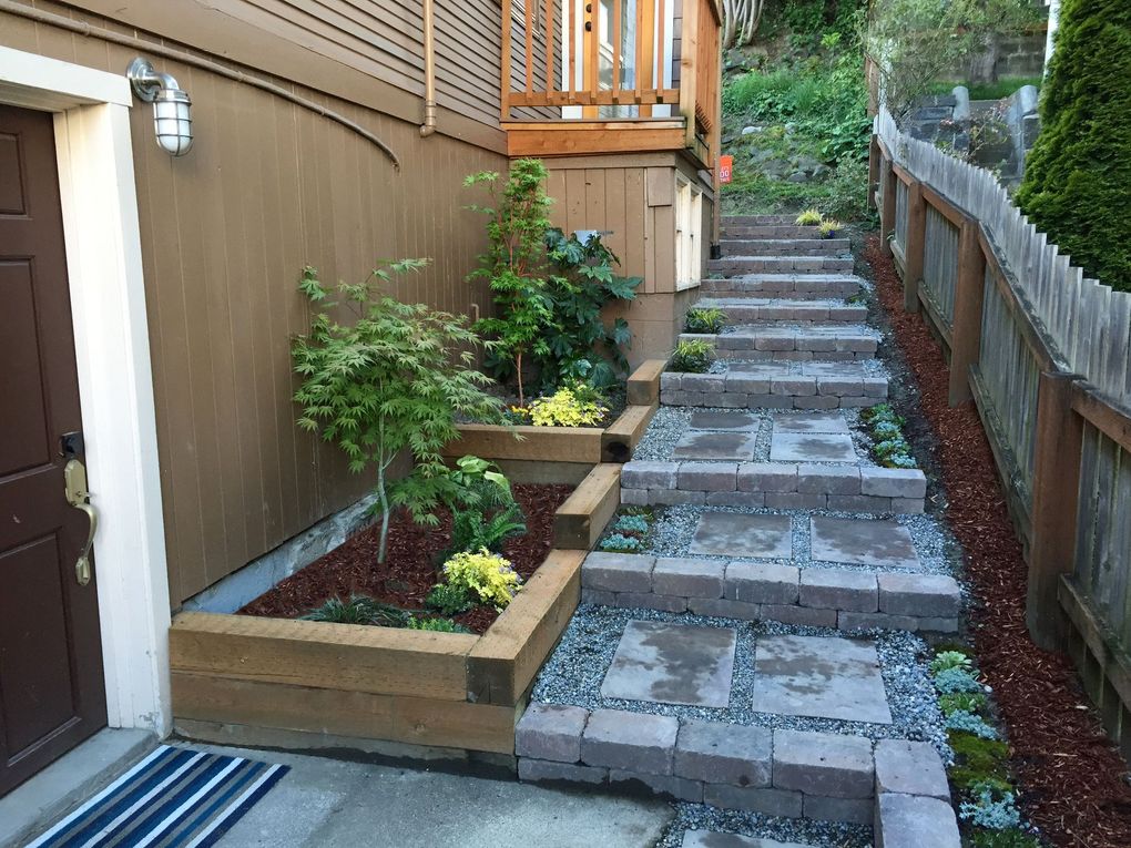 Outdoor stone staircase with wooden garden beds, mulch, and small shrubs leading up to a wooden house porch.