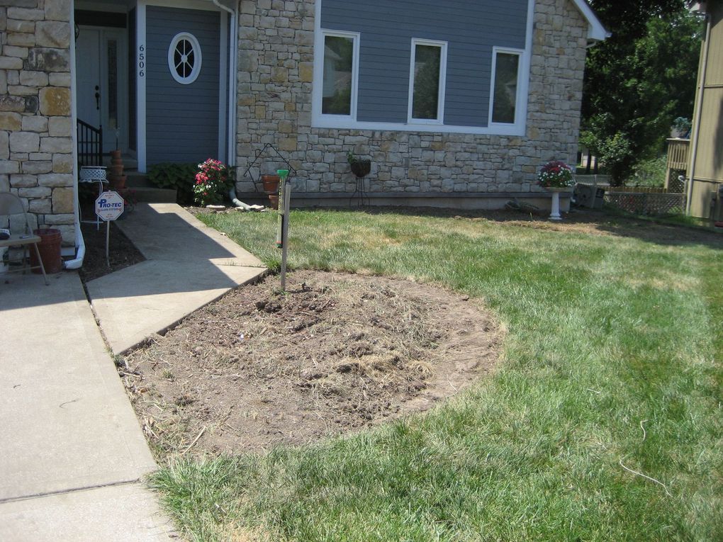 A front lawn of a stone-faced house with a circular, dirt garden bed in the foreground and a concrete walkway.