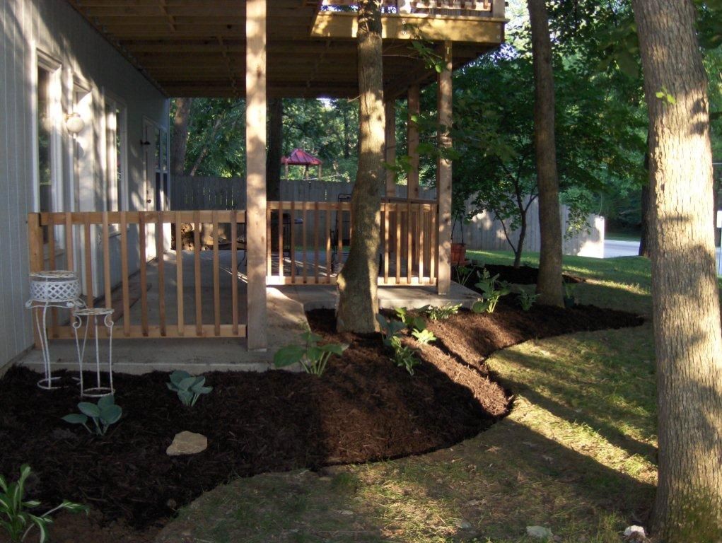 A ground-level view of a house deck with wooden railings, dark mulch landscaping, and trees in a shaded yard.