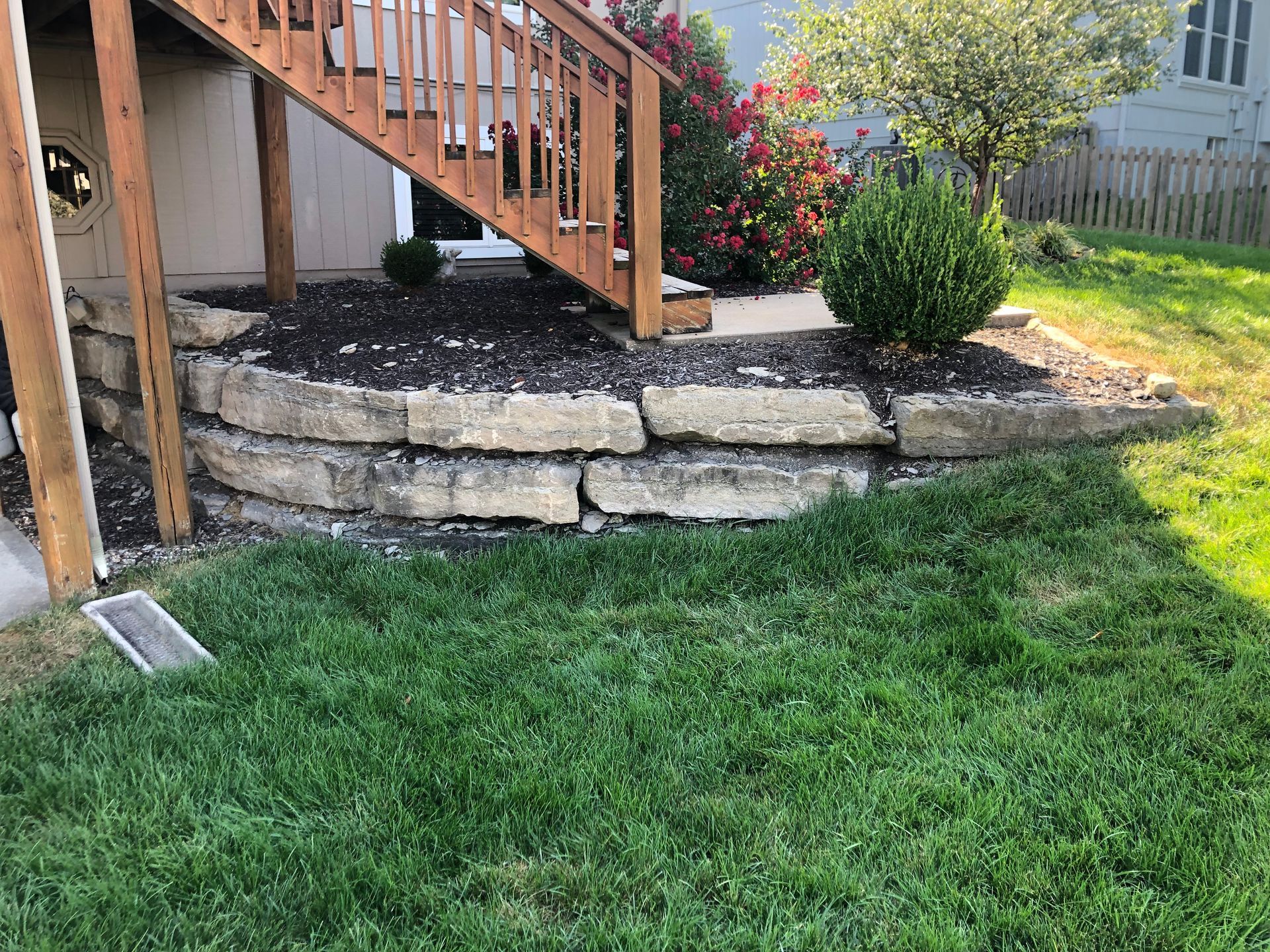 A tiered stone retaining wall beneath a wooden deck staircase in a grassy backyard.