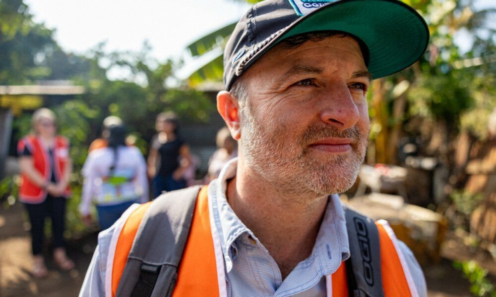 A man wearing a hat and an orange vest is standing in front of a group of people.