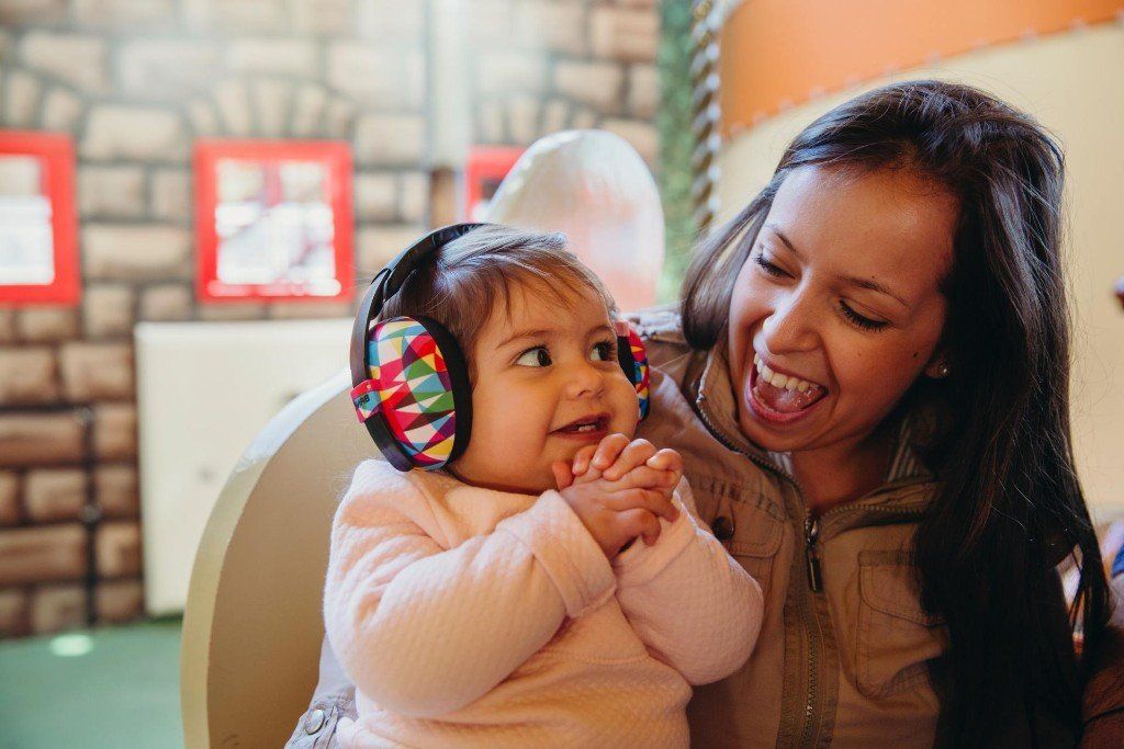 A baby wearing sound-canceling earmuffs smiles at a laughing woman, in a colorful play area.