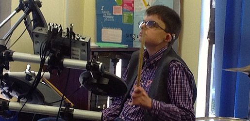 A teenage boy playing drums, wearing glasses and a hearing aid, indoor setting.