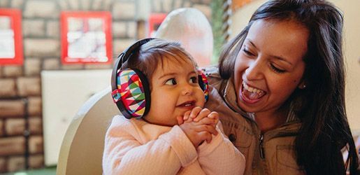 Baby wearing ear protection, smiling, sitting with a smiling woman.