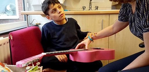 A young person is being assisted by a woman in playing a pink ukulele. They are in a room with a window.