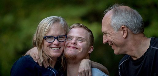 A woman, a man with Down syndrome, and a man are smiling at the camera outside.