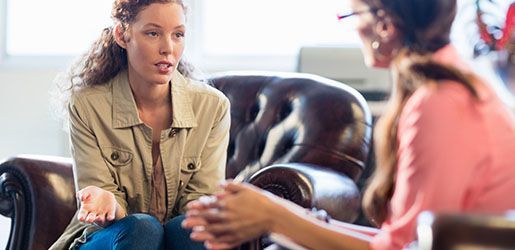 Woman talking to a therapist. The woman wears a tan jacket and jeans, gesturing while seated. The therapist is in pink.