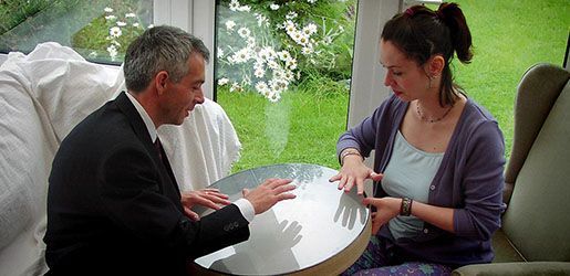 A man and woman at a round table, hands on its surface. Sunny room, flowers in background.