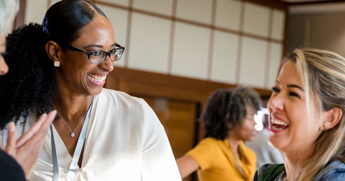 Two women smiling and talking in a well-lit room, possibly at a conference. Others in the background.