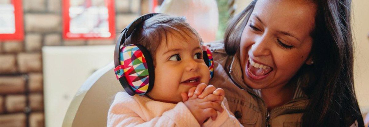 A smiling baby wearing colorful earmuffs, held by a smiling woman. Indoors, close up.