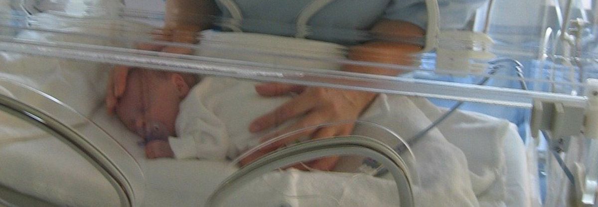 A baby inside a clear incubator; a person's hands are gently touching the baby.