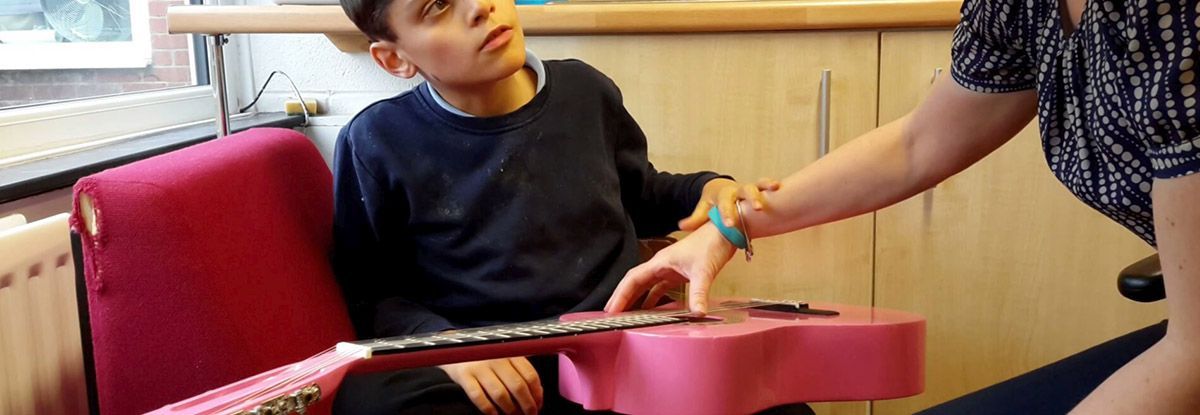 A person with assistance plays a pink ukulele. A person helps them press the strings.