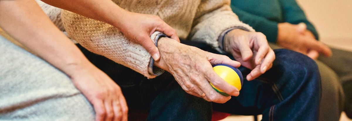 Person's hand holding a yellow ball is supported by another person's hand, possibly for physical therapy.