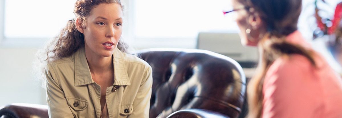 Woman in beige jacket talking to therapist, sitting in a leather chair.