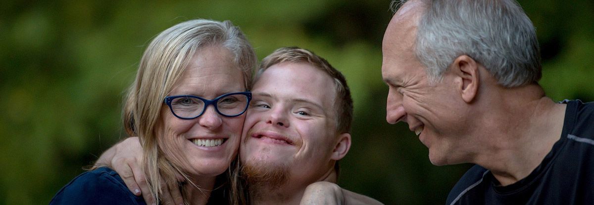 A smiling woman hugs a smiling man with Down syndrome as a man smiles nearby.