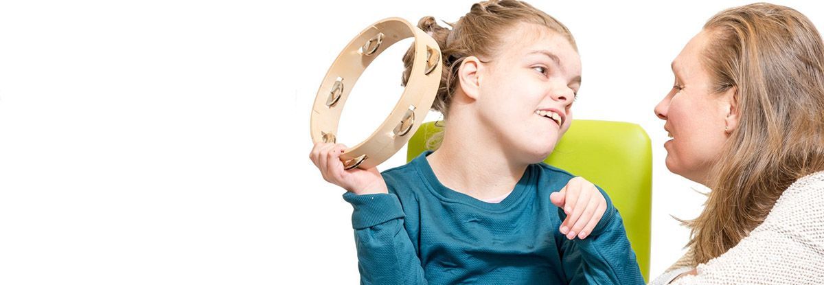 A woman and a young person with a disability are playing with a tambourine indoors, the young person holding it up.