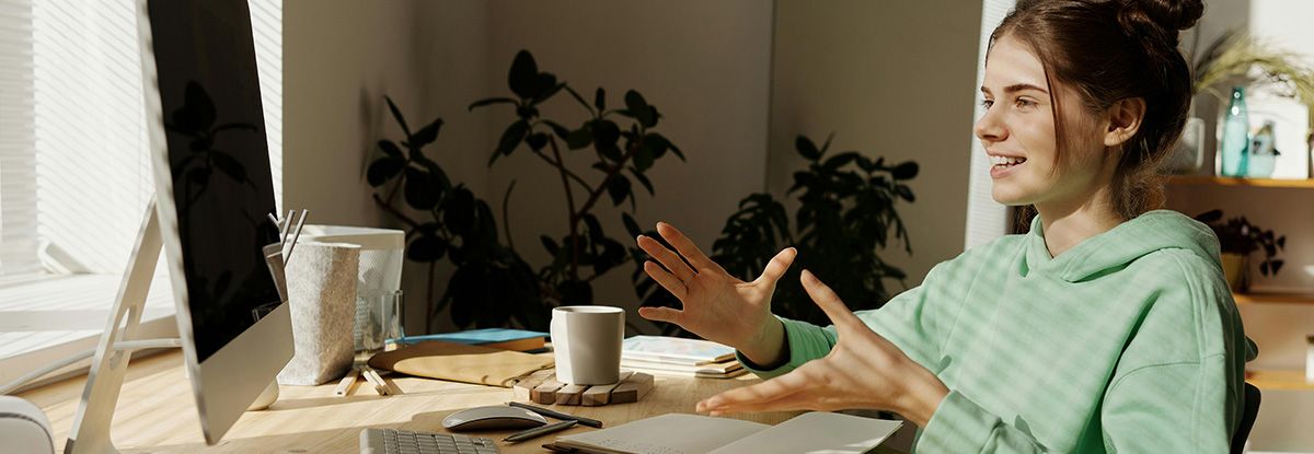 Woman with hands raised, looking at computer screen, in a home office setting. She wears a green hoodie and has a bun.