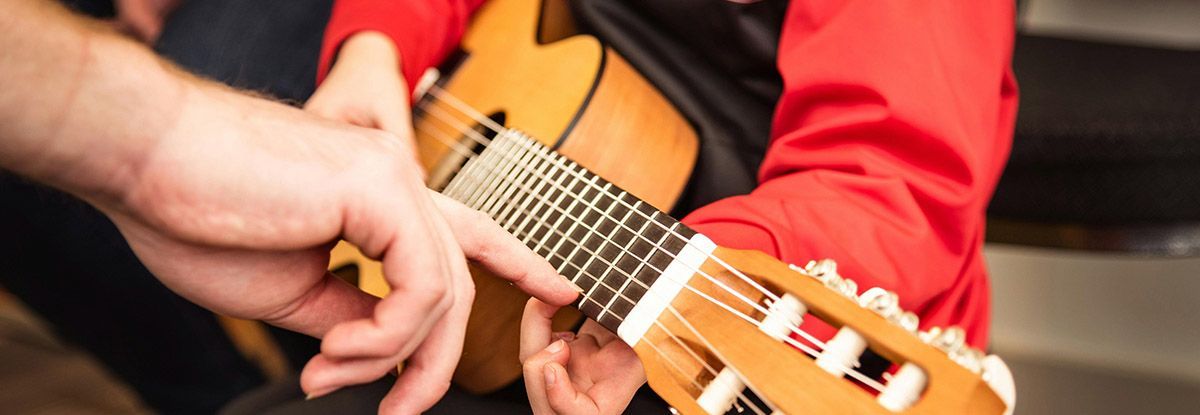 Person learning to play acoustic guitar with a hand assisting on the fretboard.