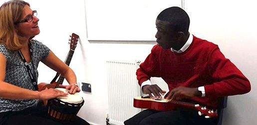A woman and a man playing music together in a white room. Woman playing a drum and man playing an ukulele.