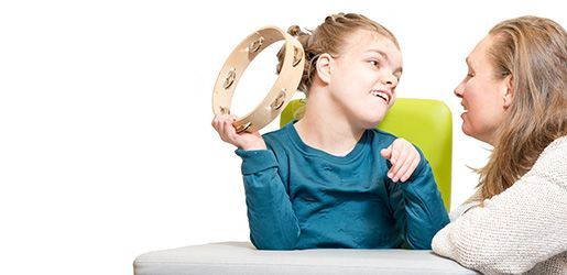 Young girl holding tambourine, looking at woman. Both are smiling, seated at a table.