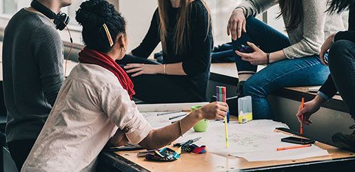 Group of students collaborating around a table with plans, using tools, and having a discussion in a bright room.
