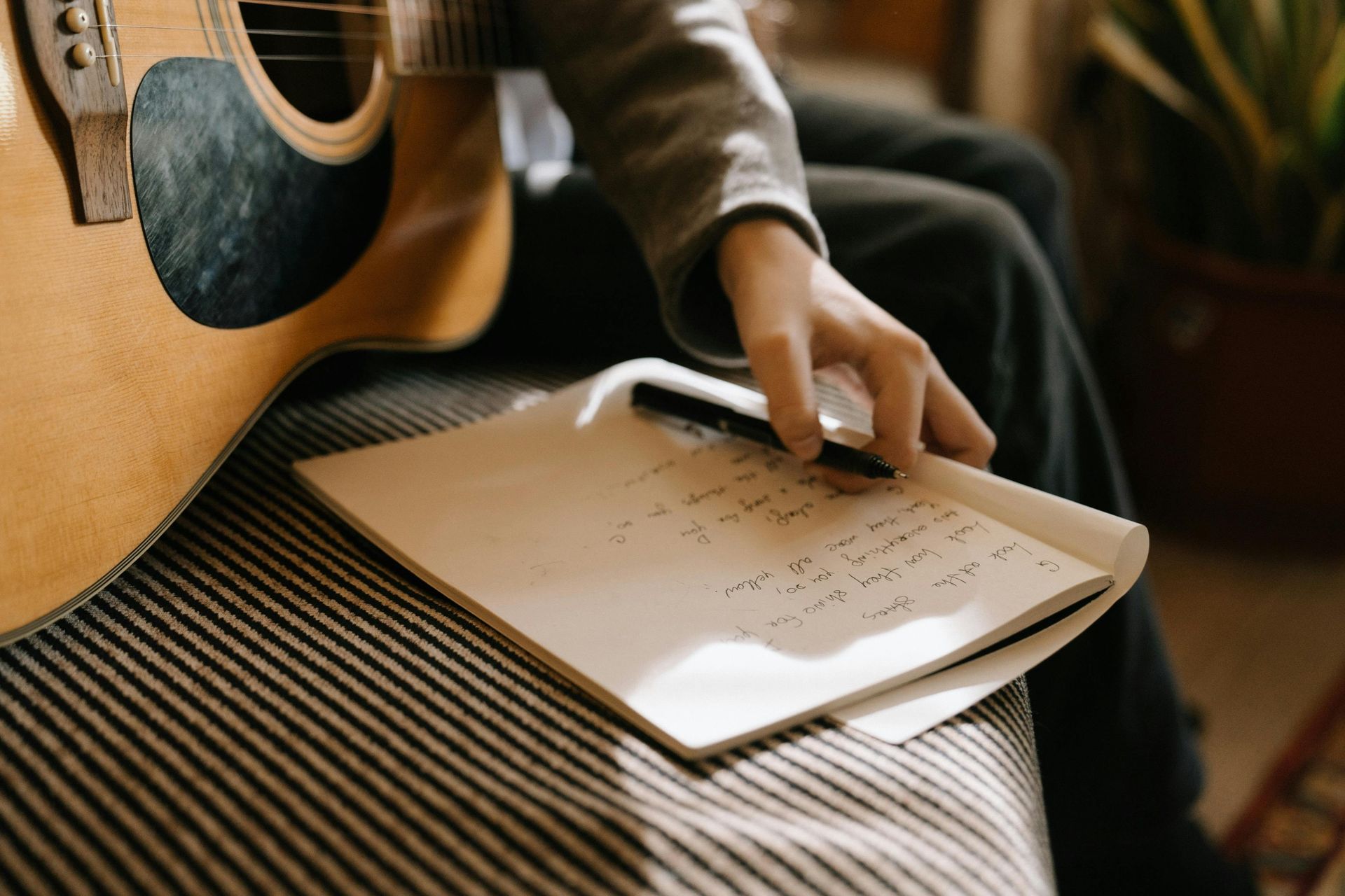 Person holding a guitar, writing in a notebook, sitting on a striped couch, natural light.