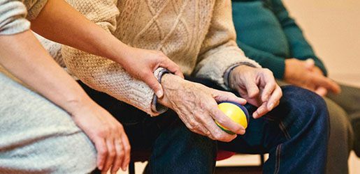 A younger hand holds an older person's wrist while the older person squeezes a yellow stress ball; seated indoors.