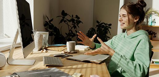 Woman in a green hoodie gesturing during a video call, seated at a desk with a computer and notebook.