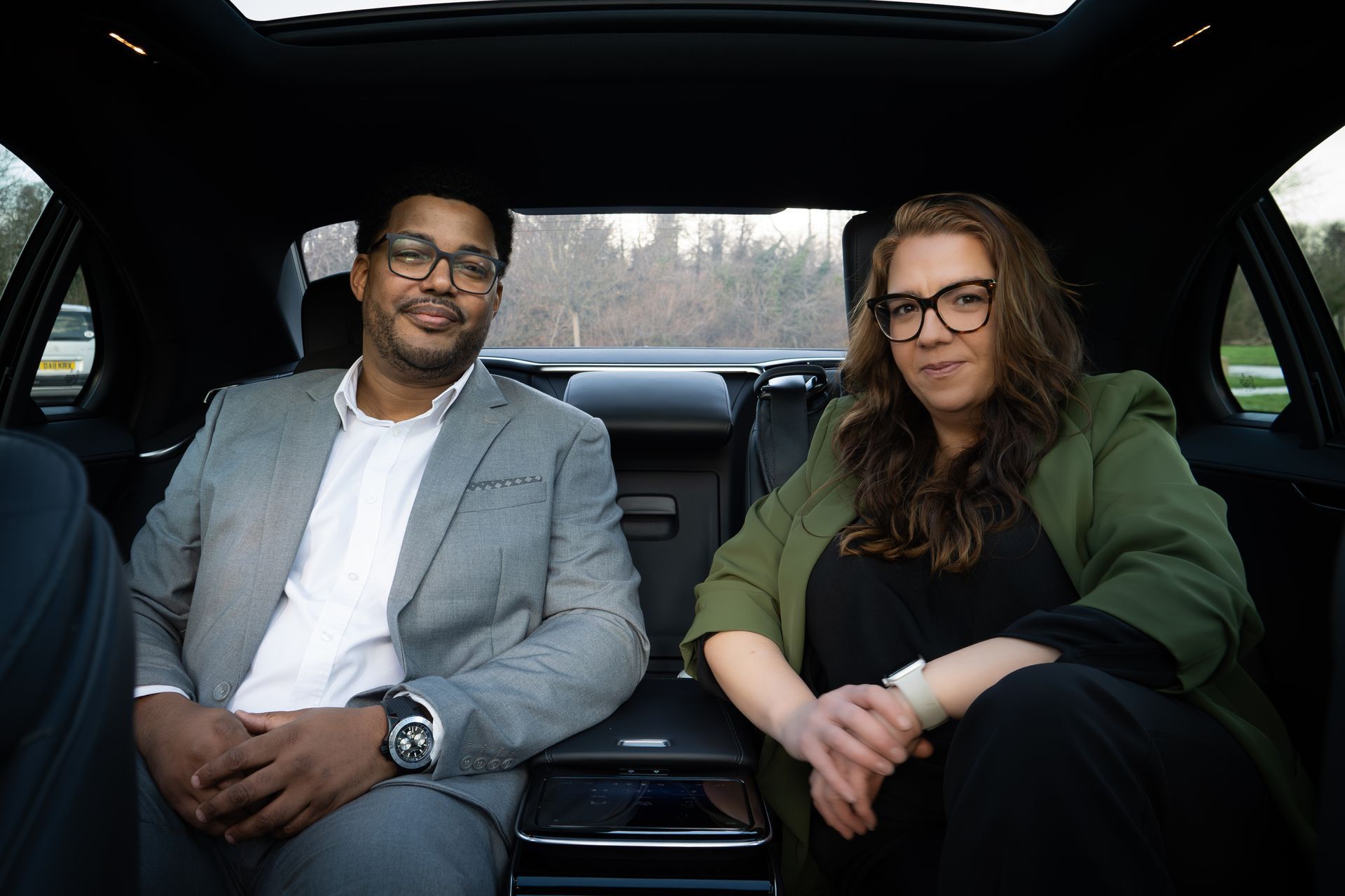 Two people sitting in the back seat of a car. Man wears a grey suit, and woman wears a green jacket.