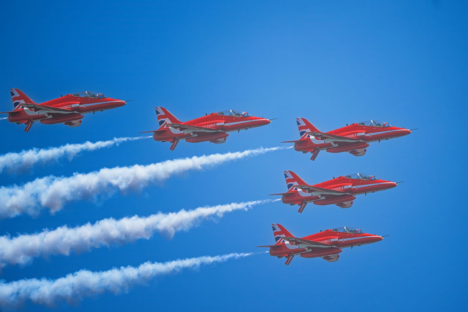 Red Hawk jets flying in formation against a blue sky, leaving white smoke trails.
