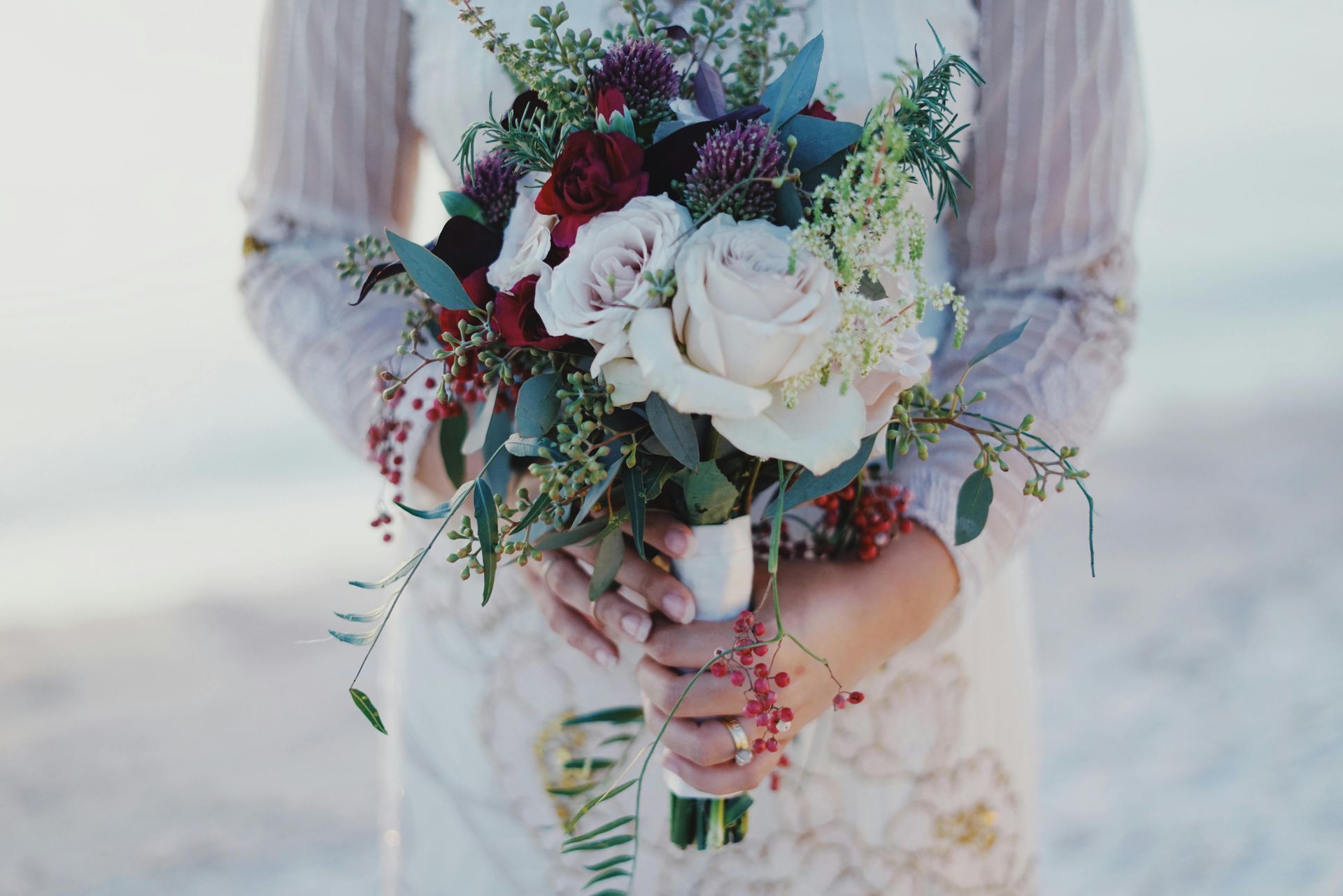 Bride holding a bouquet of roses and greenery, wearing a white dress, on a beach.