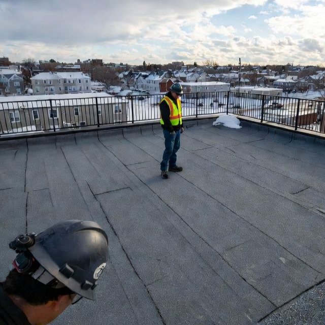 Two workers on a flat rooftop with a city view. One wears a safety vest, the other a helmet. Snowy scene.