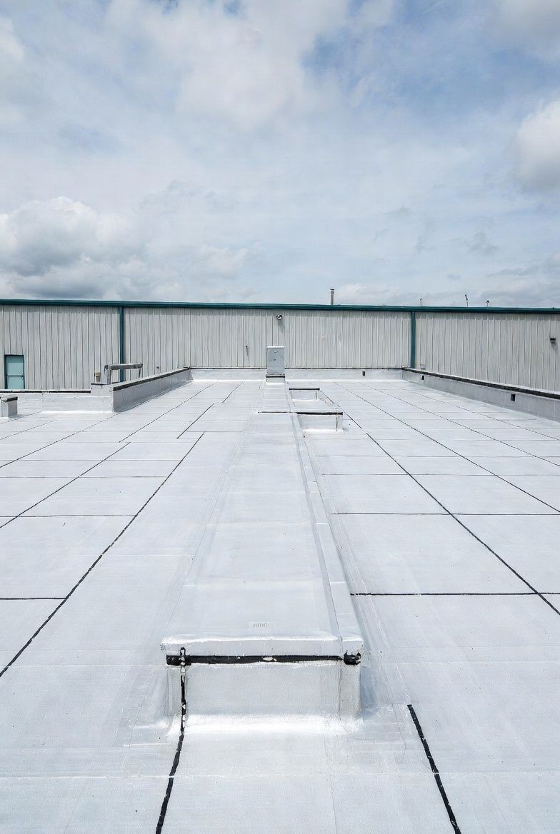 Flat, white commercial roof with rectangular panels and ventilation units. Blue sky with clouds.