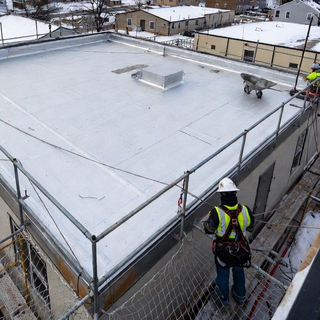 Construction workers on a flat roof with safety netting, installing silver roofing material. Snow in the background.