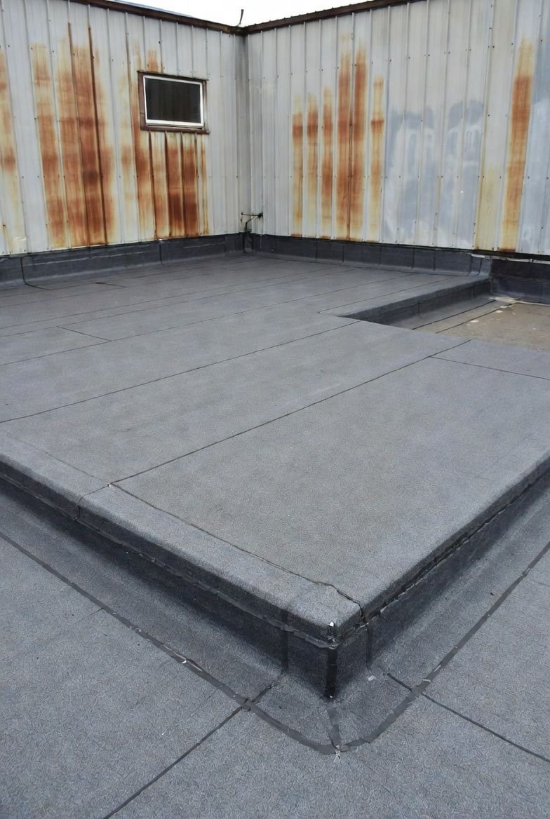 Flat, gray tar roof with dark edges and a small window on a rusty metal wall.