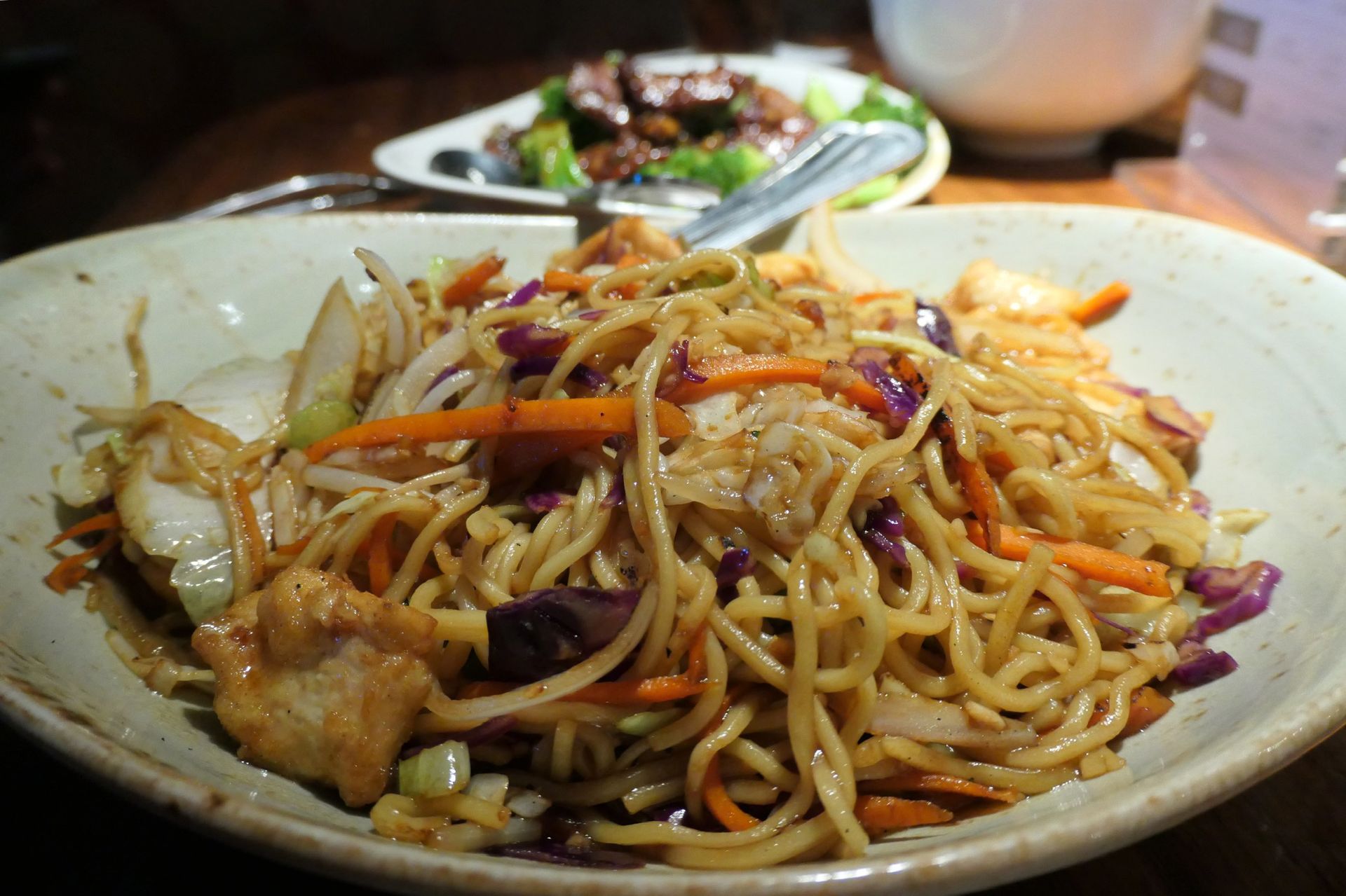 Bowl of noodles with vegetables and meat, with another dish in the blurry background.