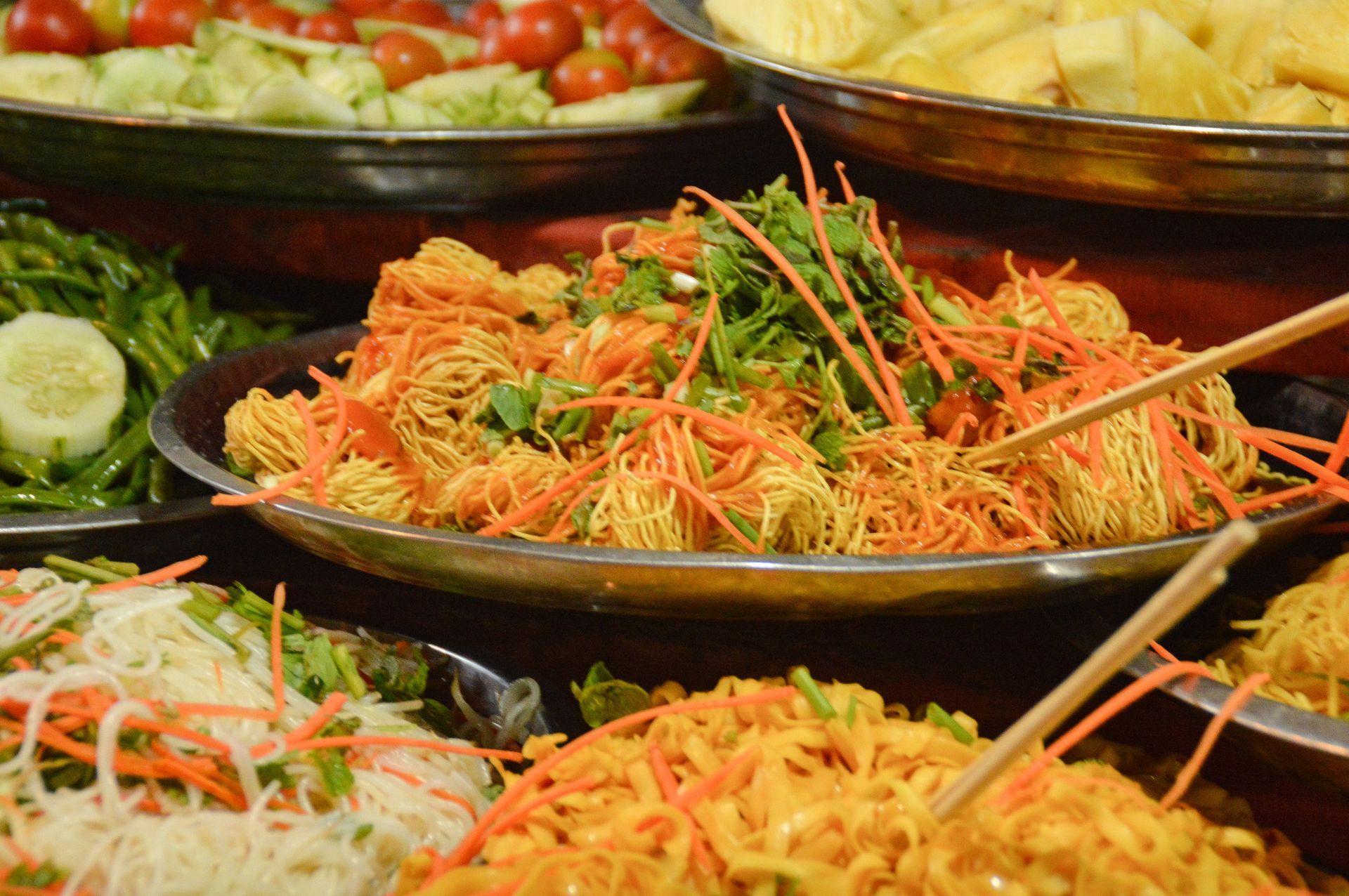 Platters of various colorful Asian dishes, including fried noodles, carrots, and garnishes, at a food market.
