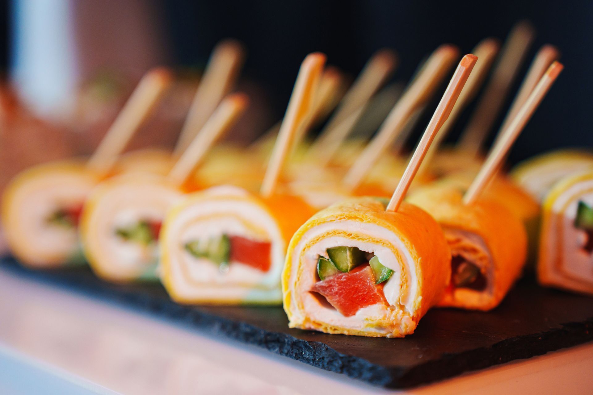 Orange crepes rolled with filling, skewered, arranged on a dark platter. Orange crepes rolled with filling, skewered, arranged on a dark platter.