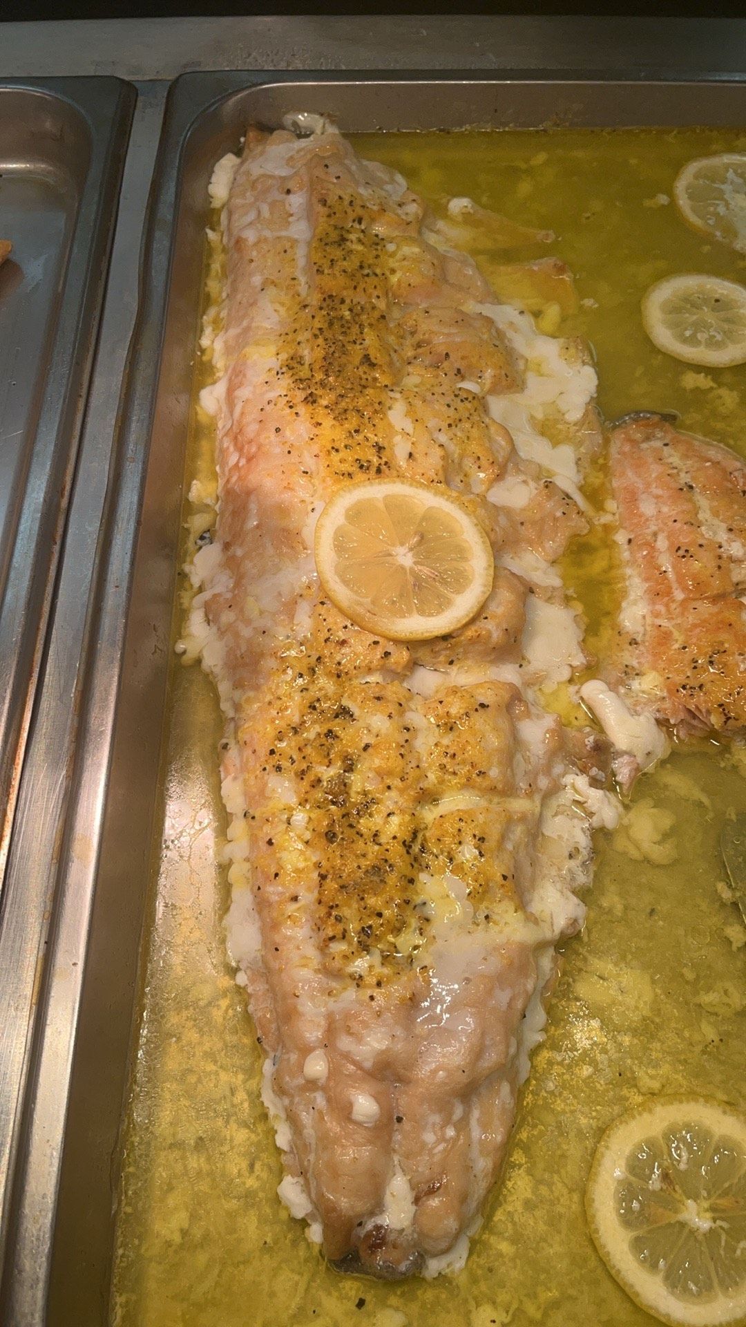 Baked fish in a tray, topped with seasoning and lemon slices, in a buttery sauce.